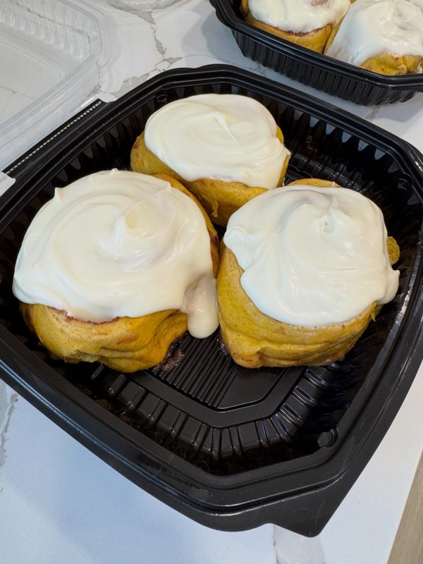 Iced pumpkin cinnamon bun in a takeout container, cream cheese icing swirled on top