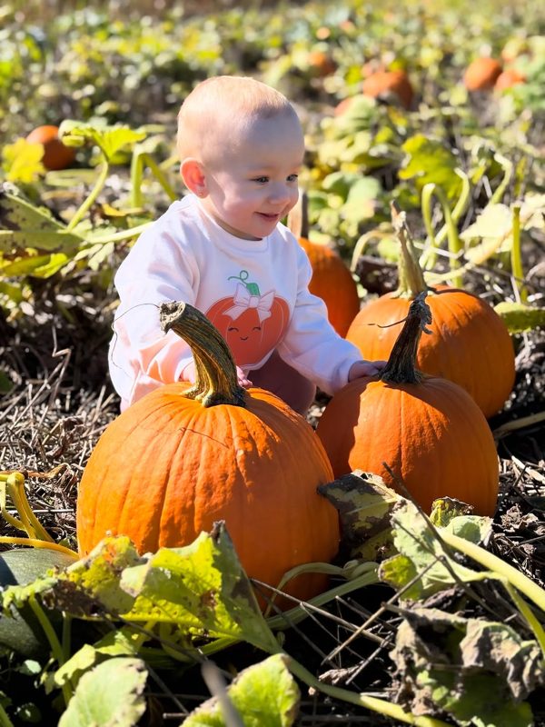 Toddler in a pumpkin-print sweatshirt sitting among pumpkins in a pumpkin patch